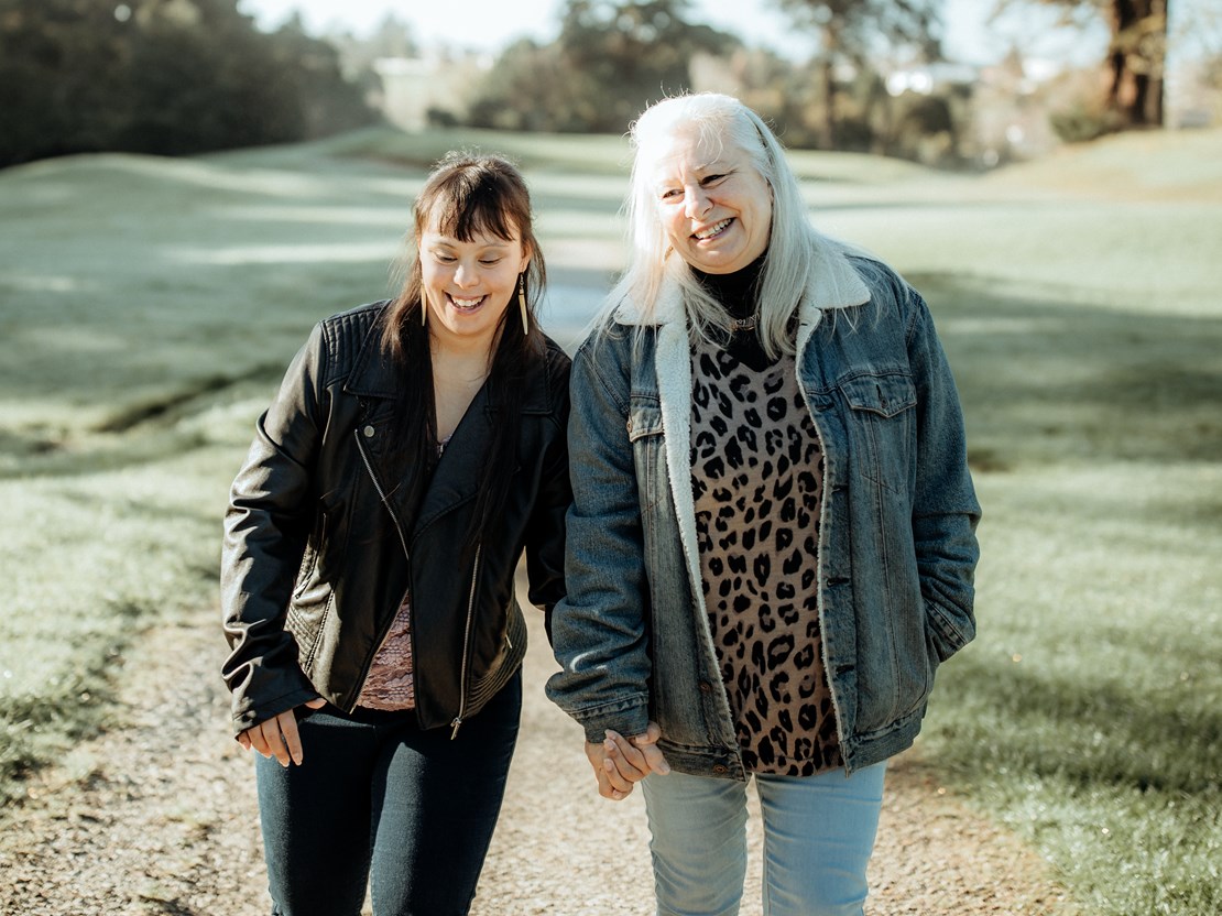 A smiling young woman with Down Syndrome and her mum walking down a path